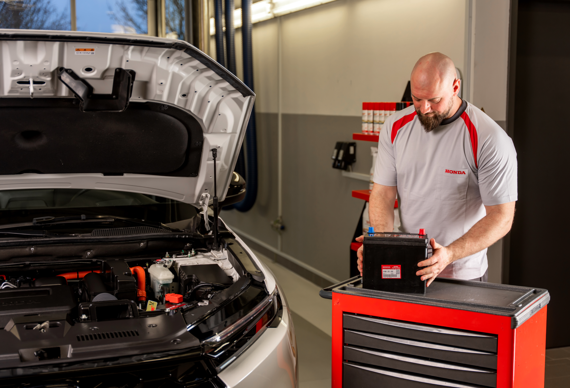 A Honda technician in a white and red uniform shirt holds a car battery next to an open hood of a Honda vehicle. A red tool cabinet is visible beside him in a garage setting.